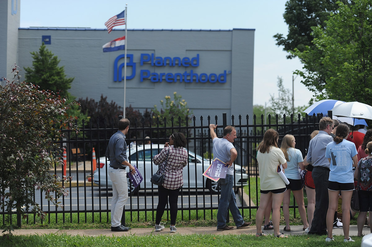 A group of people stand behind a black metal fence. In the background, a Planned Parenthood facility can be seen.