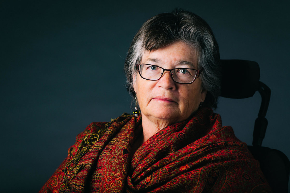 A portrait of Sandra Gail Lambert sitting in a wheelchair. She has grey hair, is wearing dark glasses, and a red pashmina scarf