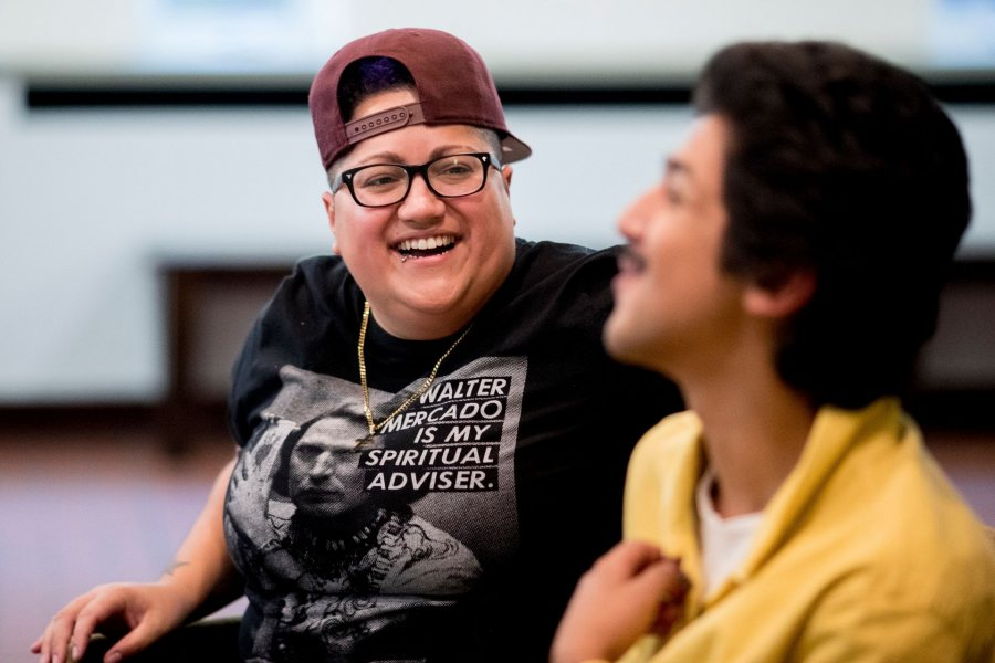 Gabby Rivera, a latinx queer woman laughs towards the camera. She is wearing a red baseball cap backwards, black glasses, and a black graphic t-shirt