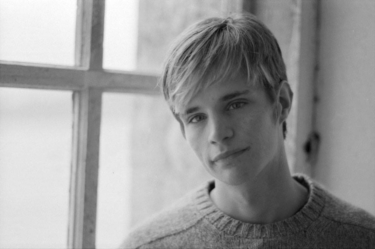 A black and white photo of Matthew Shepard. He is standing to he right of the photo with a wool sweater on. He has a shaggy blonde haircut and a soft smile