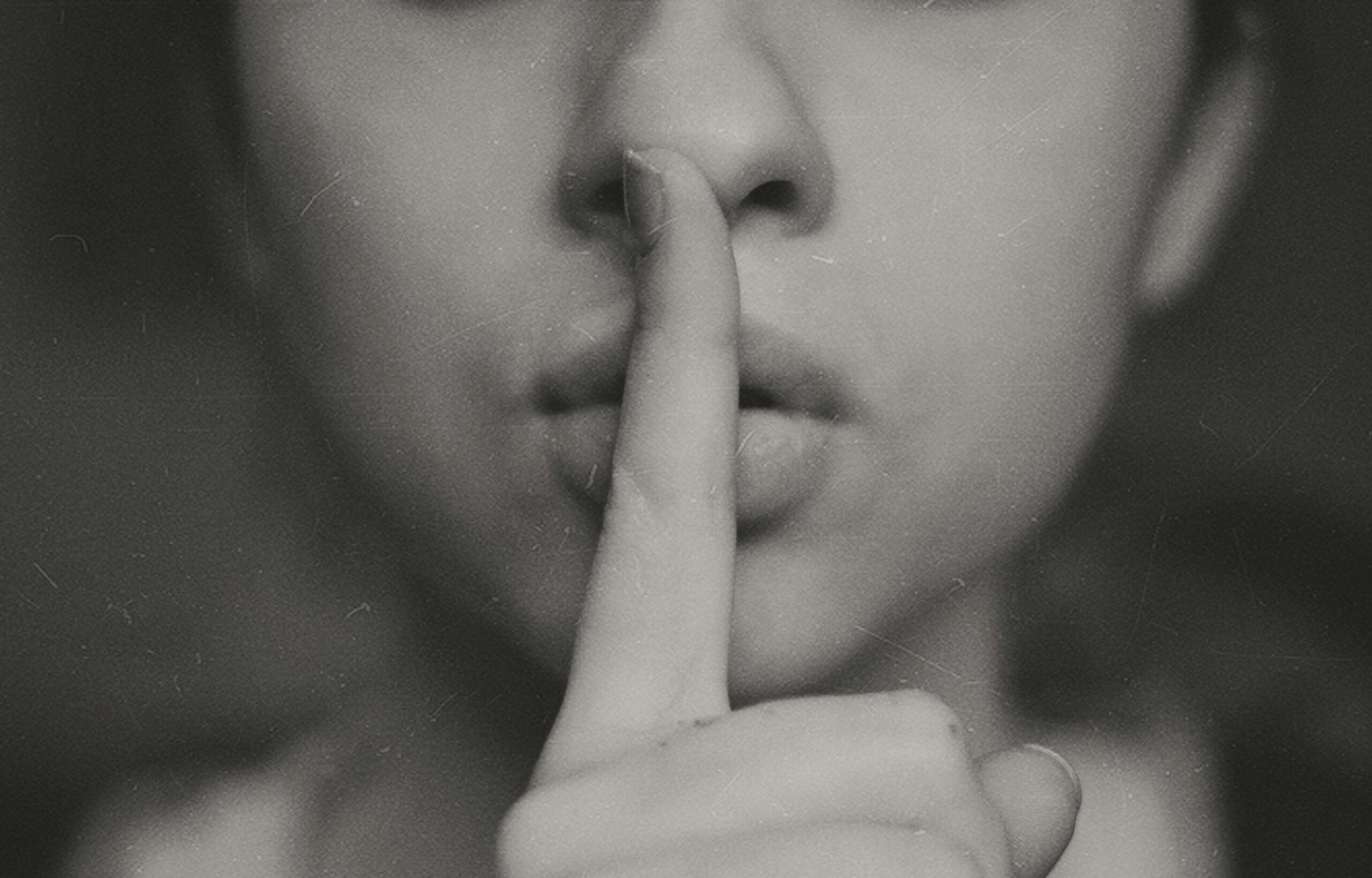 Black and white photo of a person holding their finger of their mouth to indicate the viewer should be quiet.