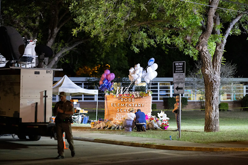 A brick sign reading "Robb Elementary School" is adorned with balloons and flowers. Two people kneel in front of the memorial.